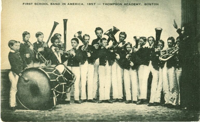 The image shows a black and white photo of the first school band in America, from Thompson Academy, Boston, in 1857. The band consists of young men in uniforms, holding various instruments such as trumpets, trombones, and drums. They are standing in front of a plain background, with a conductor on the right side.